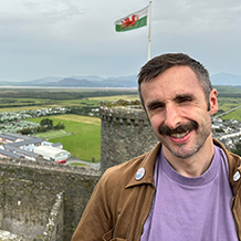 Dr Edmund Coleman-Fountain is stood smiling in the foreground. In the background there is a castle and a Welsh flag.